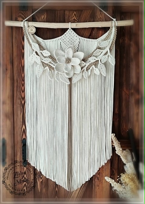 Cream-colored macrame wall hanging with fringe and leaf motifs suspended from a driftwood branch against wooden background